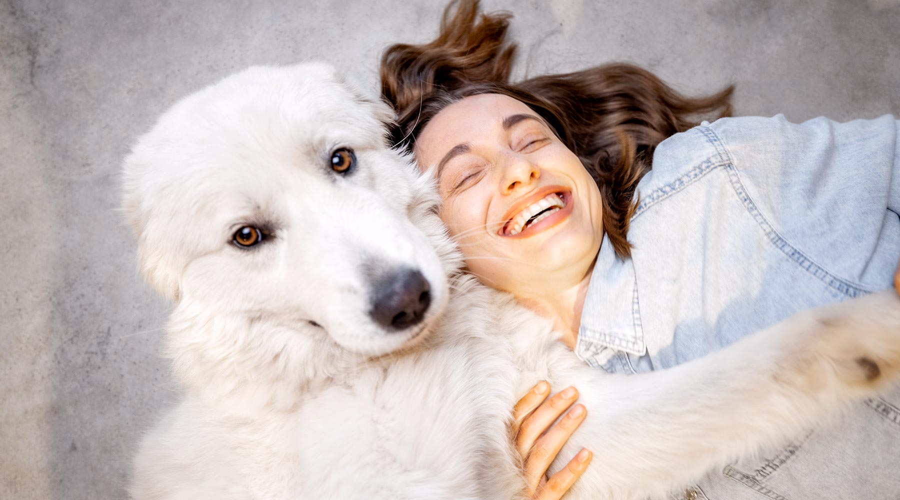 laughing-woman-lying-on-floor-with-her-fluffy-white-dog-1800x1000-1